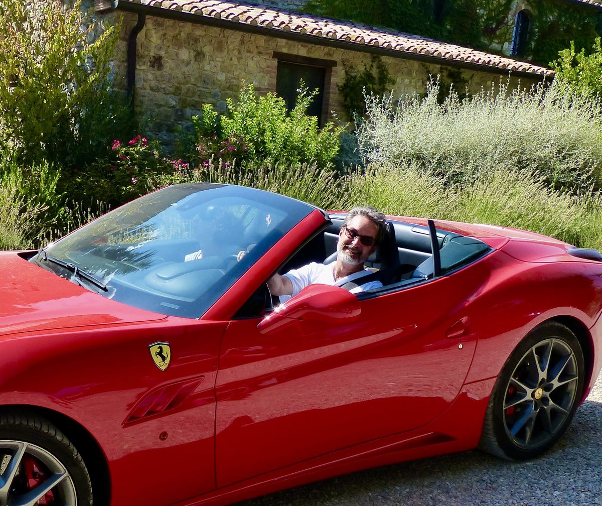 Kevin Buehler smiling from a red Ferrari convertible with the top down, a stone farmhouse and lavender behind him.