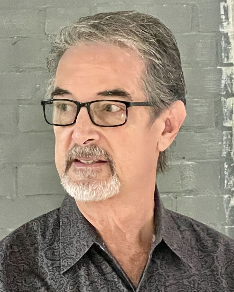 Kevin Buehler, head and shoulders, in glasses and a dark patterned shirt, in front of a gray painted brick wall.